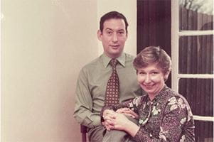 As Joy's husband sits in a chair, Joy sits beside him on the floor with her hands clasped on his knee.